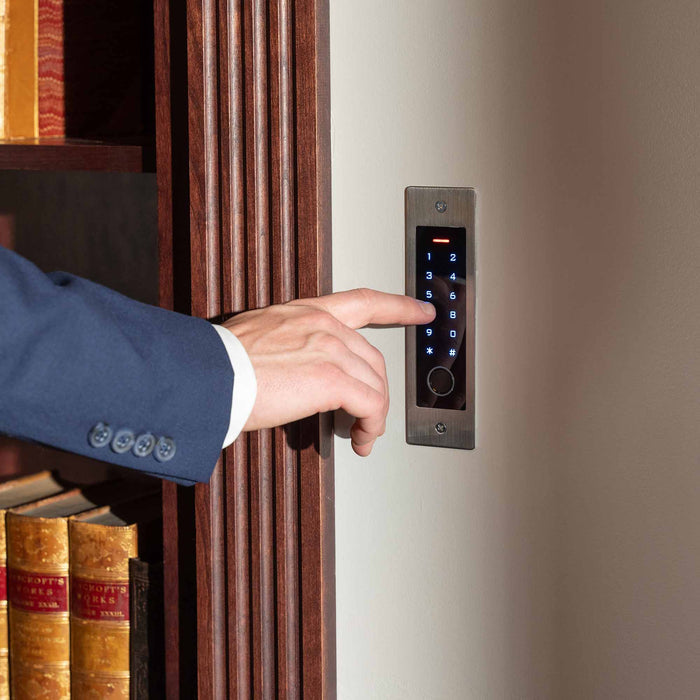 Person pressing button on sleek digital keypad lock for Murphy Door® hidden door system with wooden frame and bookshelf visible.